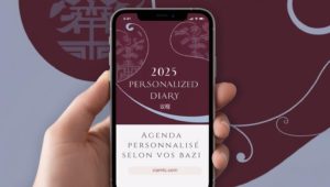 L’image montre une main tenant un téléphone intelligent affichant la couverture d’un agenda personnalisé pour 2025. L’écran du téléphone présente un design élégant aux teintes bordeaux et bleu-gris, avec des motifs asiatiques stylisés et des caractères chinois. Le texte indique : "2025 Personalized Diary" suivi du mot chinois "议程". En dessous, une section blanche affiche en français : "Agenda personnalisé selon vos Bazi", avec un lien vers "ciamtc.com". L’arrière-plan reprend les mêmes couleurs et motifs que l’écran, créant une harmonie visuelle entre le téléphone et son environnement.