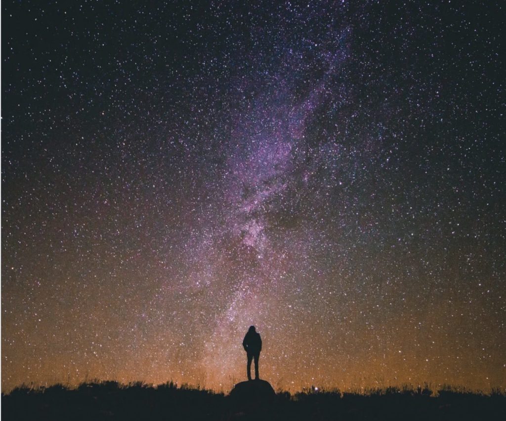 Une silhouette humaine se tient debout sur un rocher, face à un ciel nocturne rempli d'étoiles. La Voie lactée, aux teintes violettes et bleutées, s'étire en une traînée lumineuse au centre de l'image. L'horizon, légèrement orangé, crée un contraste avec l'immensité sombre du ciel. L'ensemble dégage une atmosphère de contemplation et d'émerveillement devant l'univers.