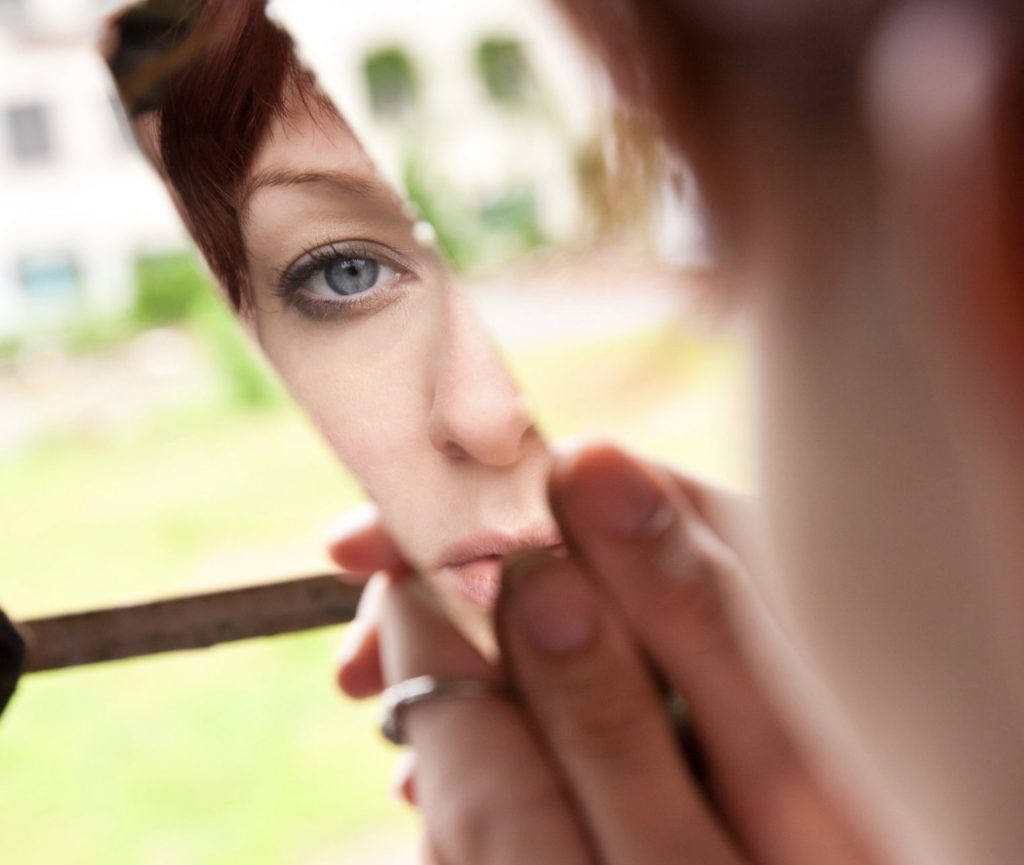 L’image montre une femme au teint clair regardant son reflet dans un morceau de miroir brisé. Son œil bleu est bien visible dans le reflet, encadré par des cheveux roux courts. Elle tient le morceau de verre avec ses doigts délicats, ornés d’une bague en argent. L’arrière-plan est flou, avec des tons verts et blancs, suggérant un environnement extérieur. La composition met en avant une ambiance introspective et fragile, accentuée par la cassure du miroir.