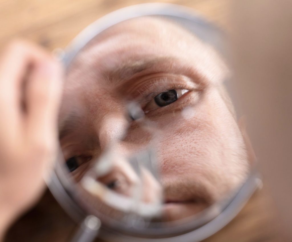 L’image montre le reflet d’un homme dans un miroir brisé. Son regard est intense, avec un œil bleu-gris bien net au centre de la composition. La fissure du miroir déforme partiellement son visage, créant une impression de fragmentation. Sa peau est légèrement texturée, avec une barbe naissante. Une main floue tient le miroir au premier plan, tandis que l’arrière-plan en bois apporte une touche chaleureuse. L’image évoque une réflexion intérieure ou une sensation de vulnérabilité.