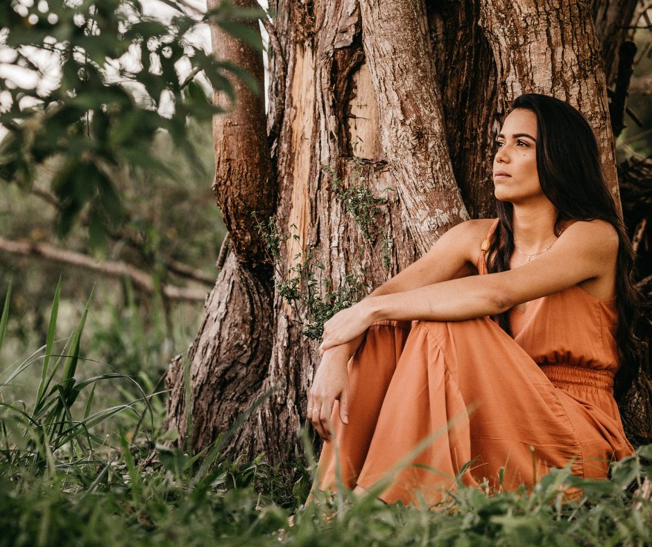 Une femme aux longs cheveux noirs et à la peau hâlée est assise adossée à un tronc d’arbre massif dans un cadre naturel verdoyant. Elle porte une robe longue de couleur terre cuite, aux fines bretelles, qui s'harmonise avec les tons chauds de l'écorce derrière elle. Son regard est tourné vers l’horizon, une expression paisible et contemplative sur le visage. L’herbe haute et les branches floues au premier plan ajoutent une ambiance douce et immersive à la scène.